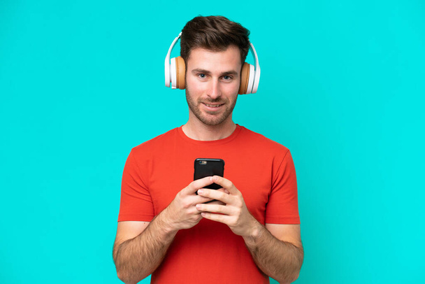 Giovane uomo caucasico isolato su sfondo blu ascoltare musica con un cellulare e guardando davanti - Foto, immagini