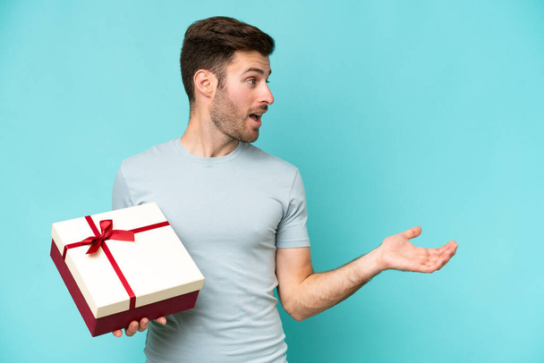 Young caucasian man holding a gift isolated on blue background with surprise facial expression - Фото, зображення
