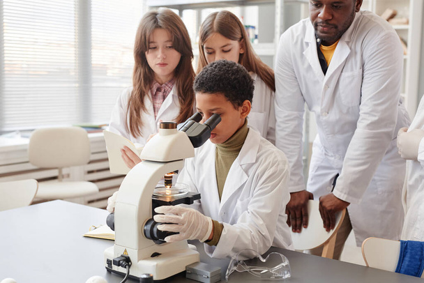 Portrét mladé černošky dívá do mikroskopu s zatímco dělá experimenty se skupinou dětí ve škole chemie laboratoř - Fotografie, Obrázek