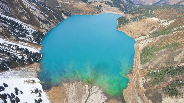A lake in the mountains with turquoise blue water. Drone view of clear water, coniferous trees and snowy mountains. People walk along the shore, low bushes grow. Big Almaty lake. Kazakhstan - Fotografie, Obrázek