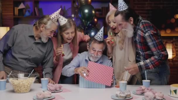 笑顔のシニアおじいちゃんは、お祝いの誕生日キャップ帽子をかぶって、リボンでギフトボックスを開きます退職ホームで他のシニアと一緒にお祝いします。誕生日パーティーギフト - 映像、動画
