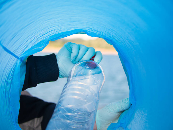 Λεπτομέρεια ενός χεριού που βάζει ένα πλαστικό μπουκάλι μέσα σε μια μπλε σακούλα σκουπιδιών που φαίνεται μέσα από την τσάντα. Ανακύκλωση εννοιών, σκουπίδια, πλαστικά, ρύπανση, περιβάλλον. - Φωτογραφία, εικόνα