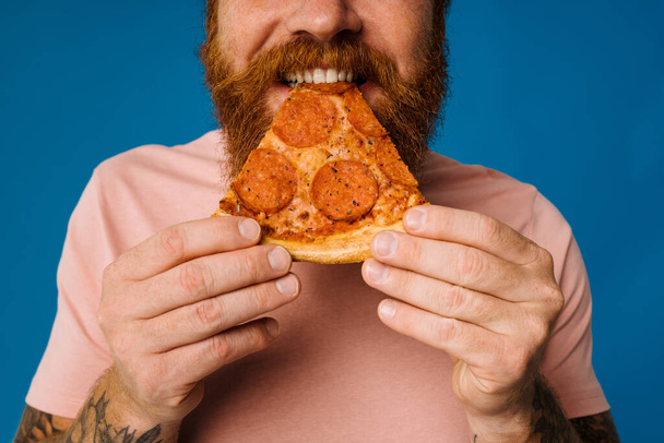 Bearded happy man eating pizza while standing isolated over blue background - Foto, Imagen