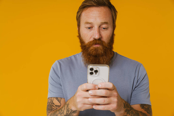 Bearded white man holding cellphone while standing isolated over yellow background - Fotoğraf, Görsel