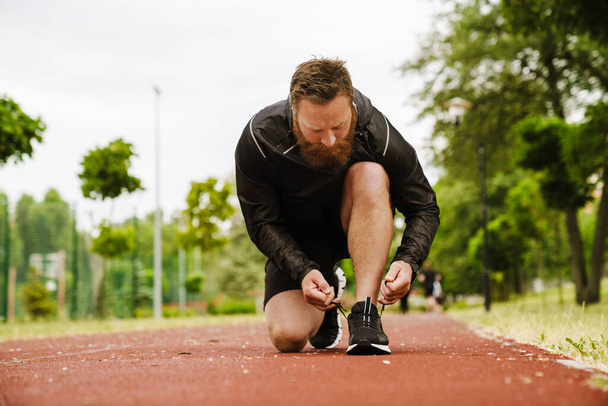 Adult handsome bearded redhead man tying his shoelaces looking downward , while standing on his knee on running track outdoors - Fotografie, Obrázek