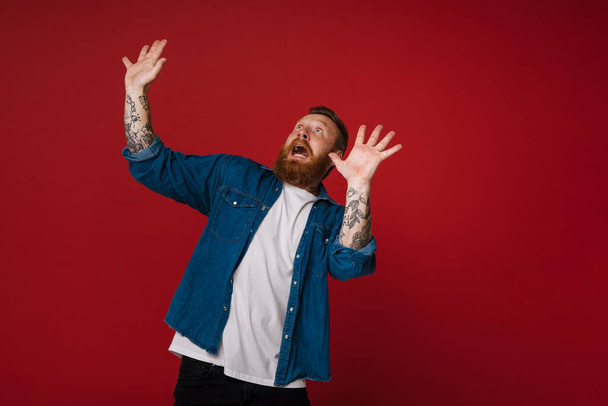 Adult bearded tattooed handsome scared redhead man with raised hands and opened mouth looking aside, while standing over isolated red background - Фото, зображення