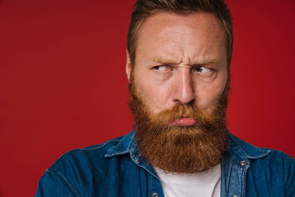 Portrait of adult bearded tattooed handsome frowning redhead man looking aside , while standing over isolated red background - Fotografie, Obrázek