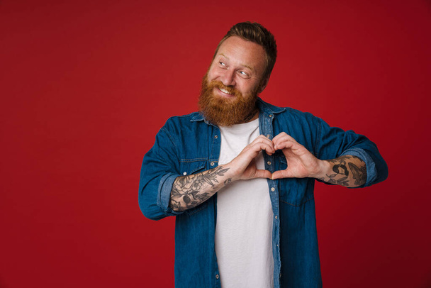 Adult redhead bearded tattooed smiling man in t-shirt doing heart gesture wih fingers expressing tender emotion and looking aside , while standing over isolated red background - Photo, Image