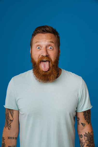 Portrait of adult bearded tattooed handsome stylish manwith wide opened eyes showing tongue and looking at camera, while standing over isolated blue background - Valokuva, kuva
