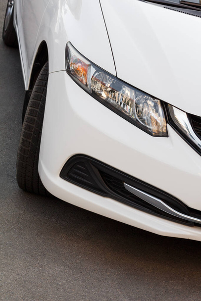 Modern white car parked on asphalt. Close-up front view with headlight, bumper and wheel - 写真・画像