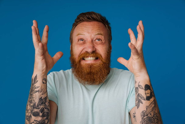 Adult bearded tattooed handsome stylish stressed man with clenched teeth raised hands and splayed fingers looking upwards, while standing over isolated blue background - Photo, Image