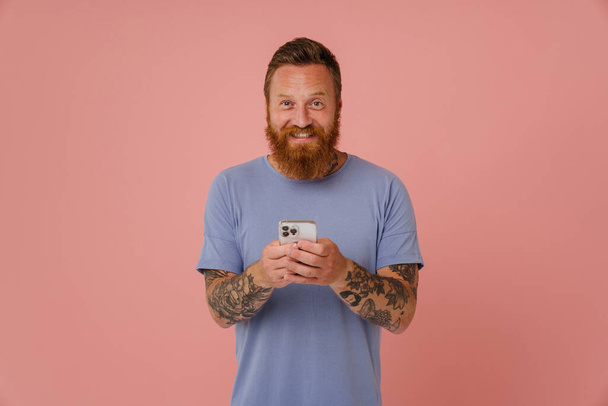 Portrait of adult bearded tattooed handsome smiling man holding phone and looking at camera, while standing over isolated pink background - Photo, image