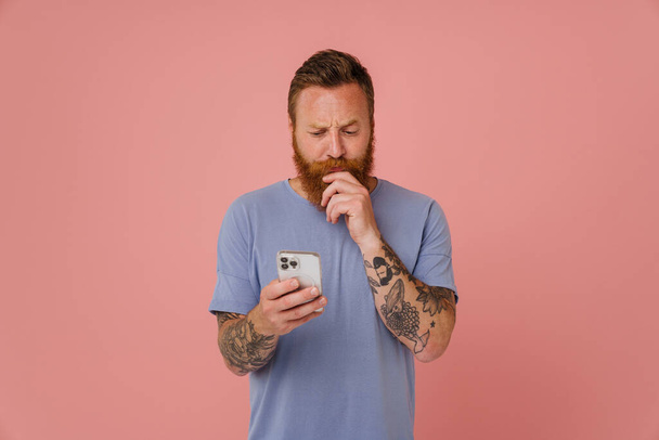 Portrait of adult bearded tattooed handsome thoughtful man looking at his phone and holding his beard, while standing over isolated pink background - Фото, зображення