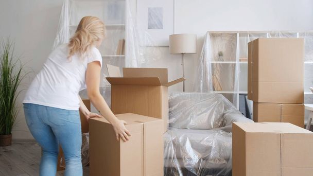 Home relocation. Moving day. Removal service. Unrecognizable woman carrying carton boxes in light modern apartment with covered furniture. - Fotoğraf, Görsel