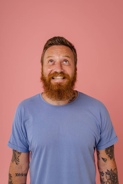 Adult handsome bearded tattooed smiling redhead man in blue t-shirt looking upwards, while standing over isolated pink background - Foto, Bild
