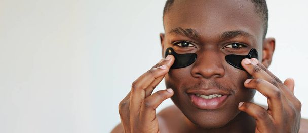 Eye patches. Facial treatment. Anti-aging skincare. Closeup portrait of guy applying black moisturizing face skin gel pads cosmetic product on light empty space background. - Fotó, kép