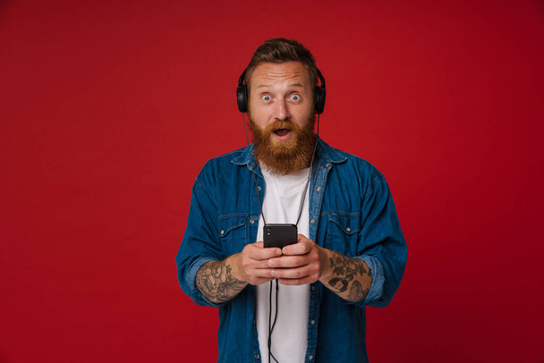 Ginger surprise man in headphones listening music and using cellphone isolated over red background - Φωτογραφία, εικόνα
