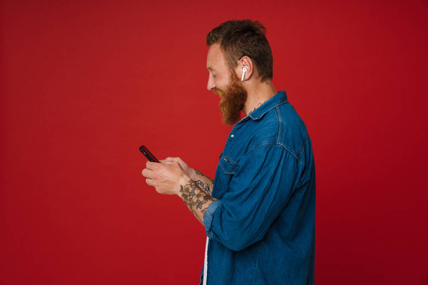 Ginger excited man in earphone listening music and using cellphone isolated over red background - Фото, зображення