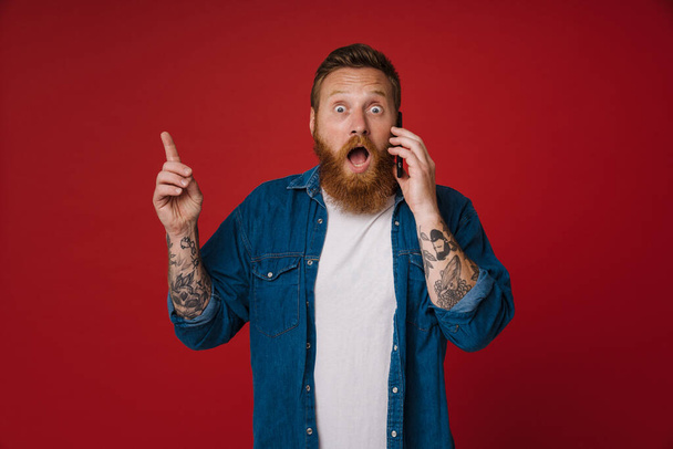 Ginger excited man wearing denim shirt talking on cellphone and gesturing isolated over red background - Photo, image