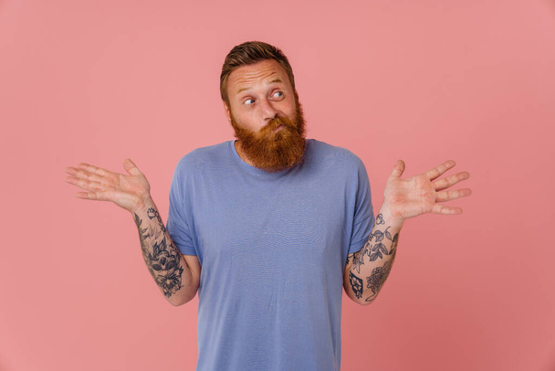 Ginger perplexed man with beard gesturing and looking aside isolated over pink background - Фото, зображення