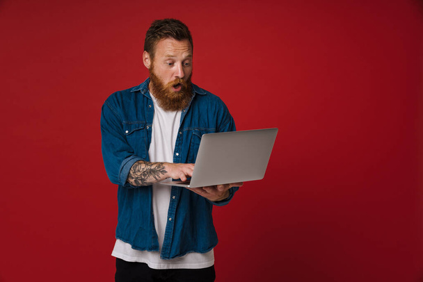 Ginger shocked man wearing denim shirt using laptop isolated over red background - Foto, Bild