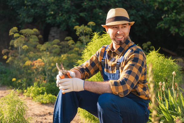 Portrait of bearded, handsome happy gardener in uniform and a straw hat sits and relax at his kitchen-garden, holding a pruner. In the background is a backyard and a garden. The concept of gardening and spring. - Photo, Image