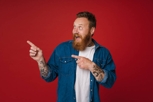 Ginger excited man with tattoo gesturing and pointing fingers aside isolated over red background - Fotoğraf, Görsel