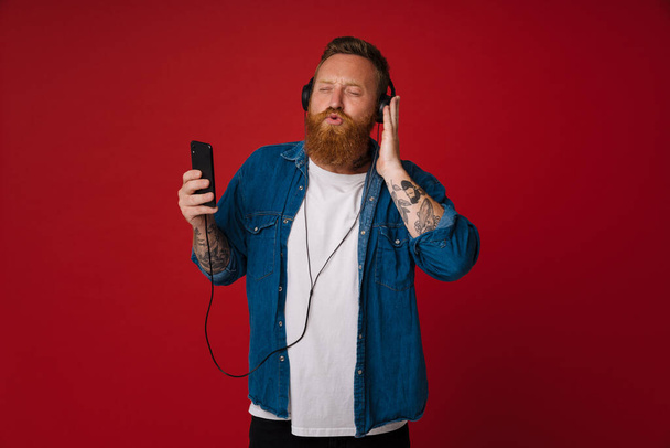 Ginger white man singing and dancing while listening music isolated over red background - Foto, imagen