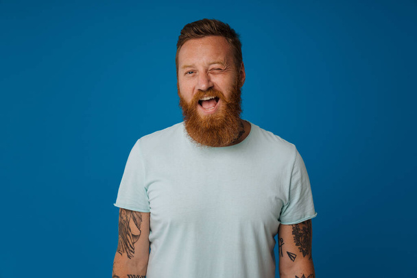 Ginger excited man with tattoo winking and smiling at camera isolated over blue background - Fotografie, Obrázek