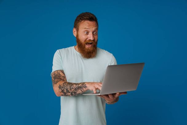 Ginger excited man with tattoo screaming and working with laptop isolated over blue background - Fotó, kép