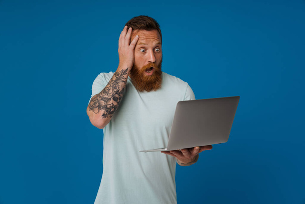 Ginger shocked man expressing surprise while working with laptop isolated over blue background - Foto, afbeelding