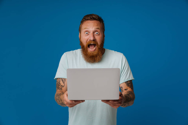 Ginger excited man with tattoo screaming and working with laptop isolated over blue background - Foto, imagen