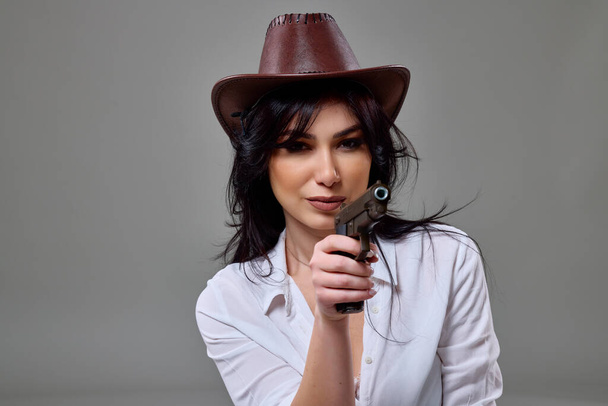 Портрет красивой женщины в ковбойской шляпе с пистолетом в руке, позирующей в студии на сером фоне - Фото, изображение