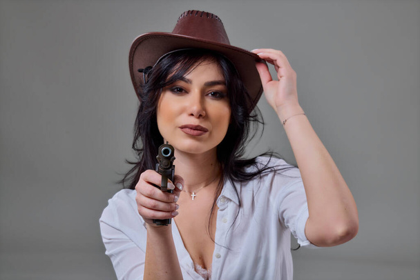 Egy gyönyörű nő portréja cowboy kalapban és pisztollyal a kezében, amint a stúdióban pózol szürke alapon. - Fotó, kép