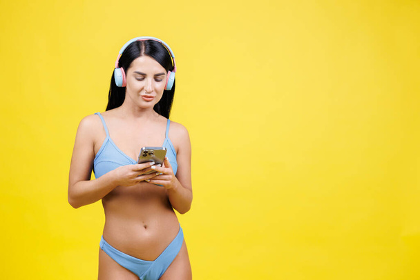 Glückliche junge Frau mit schlankem Körper trägt einen blauen Badeanzug, hört Musik über Kopfhörer und hält ihr Handy isoliert in den Händen auf gelbem Wandhintergrund. Sommerruhe-Konzept. - Foto, Bild