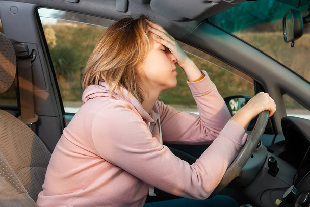 Młoda blond nerwowa kobieta siedzi za kierownicą lewostronnego samochodu i trzyma jej głowę. Widok z boku. Grzywna za naruszenie przepisów ruchu drogowego. - Zdjęcie, obraz