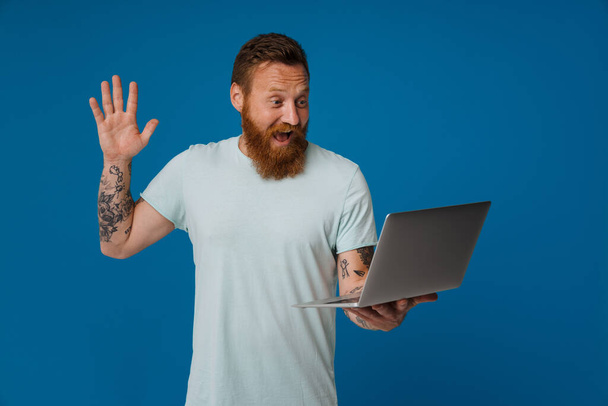 Ginger izgatott férfi mosolyog és gesztikulál, miközben dolgozik laptop elszigetelt kék háttér - Fotó, kép