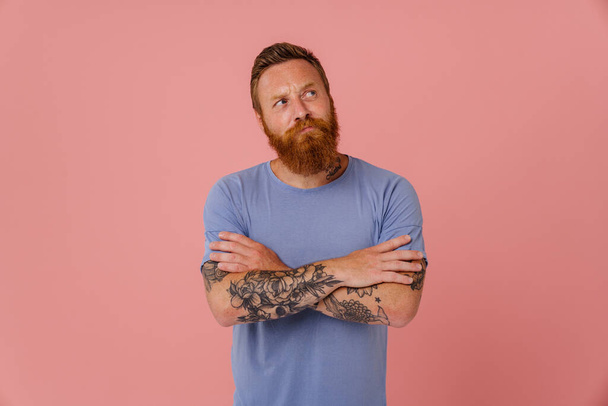 Ginger puzzled man with beard frowning and looking aside isolated over pink background - Foto, Imagen