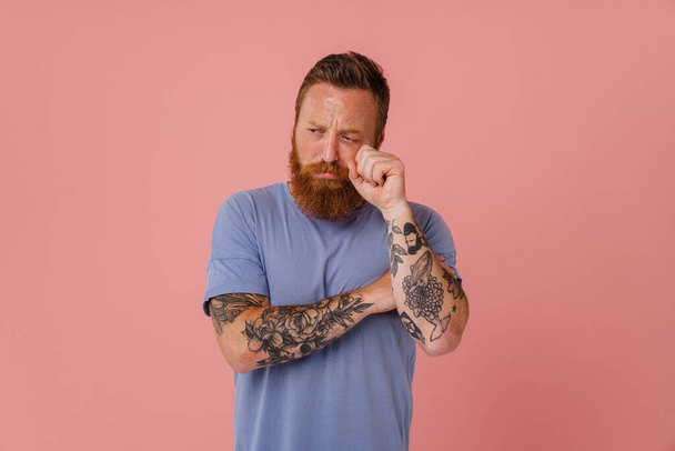 Ginger sad man with beard crying while wiping his tears isolated over pink background - 写真・画像