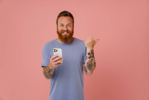 Ginger white man using cellphone and pointing finger aside isolated over pink background - Foto, Imagem