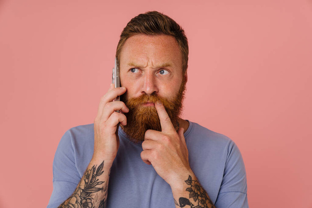 Ginger white man talking on cellphone and frowning isolated over pink background - Foto, Imagem