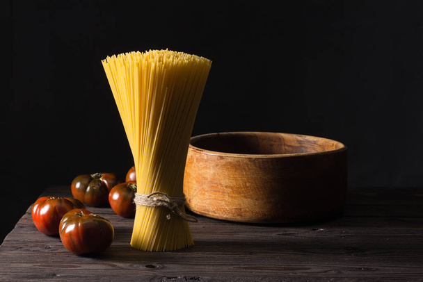Some tomatoes and a bunch of spaghetti arranged vertically on an antique table. Dark background. - Foto, Imagem
