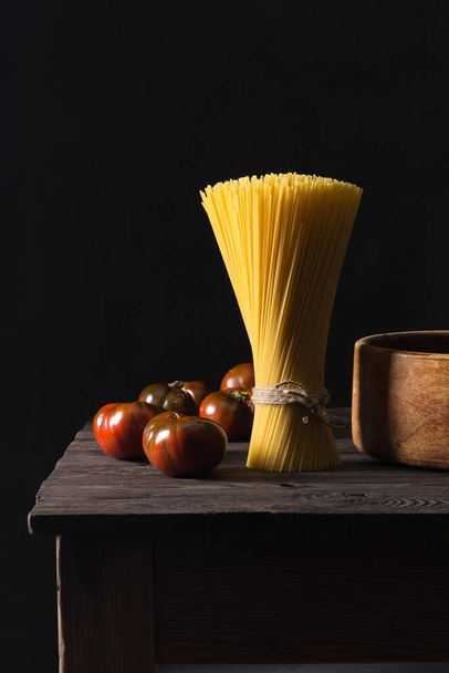 Some tomatoes and a bunch of spaghetti arranged vertically on an antique table. Dark background. - Фото, изображение
