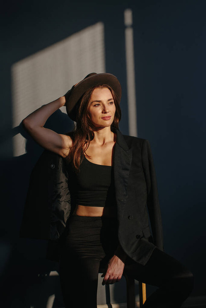 Divatportré egy gyönyörű lányról fekete öltönyben. Egy gyönyörű barna pózol a háttérben egy szürke fal egy fekete kabát és kalap, fény az ablakból. - Fotó, kép