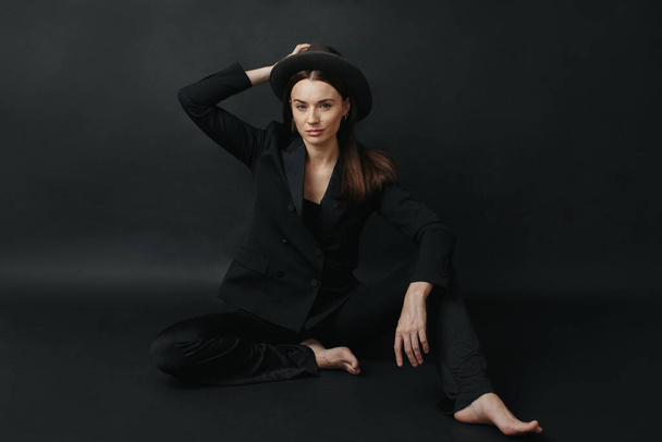 Portrait of a modern stylish woman. A brunette in a suit and hat poses barefoot on a black background. - Фото, изображение
