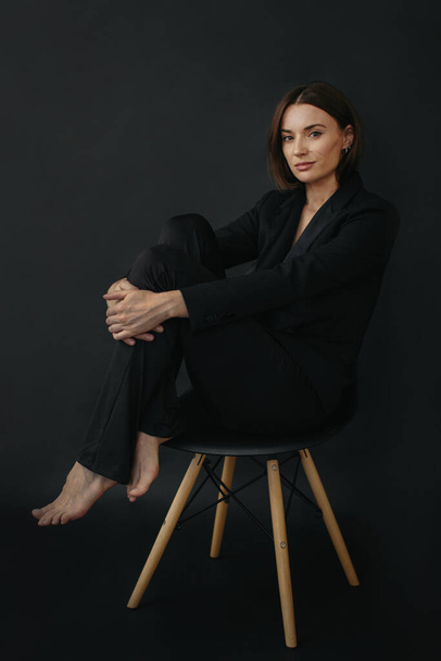 Портрет современной стильной женщины. Красивая брюнетка позирует на стуле на черном фоне. - Фото, изображение