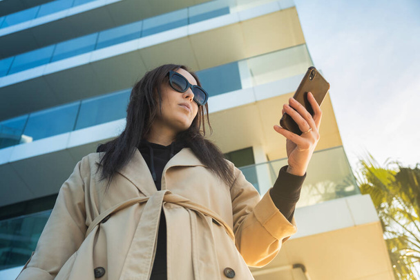 Muotokuva tyylikäs nainen älypuhelimella aurinkoisena päivänä katukaupungissa, jossa on toimistorakennukset ja auringonpurkaus taustalla. Naispuolinen johtaja käyttää matkapuhelinta ulkona - Valokuva, kuva