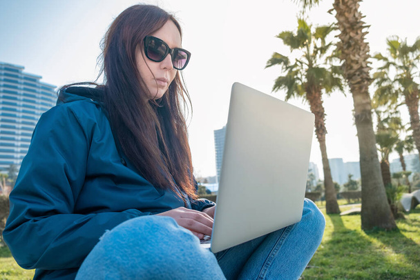 Молодая женщина фрилансер или цифровой кочевник, работающий удаленно с помощью ноутбука для общения, чата онлайн сидя в парке. Женщина-фрилансер работает на открытом воздухе за компьютером - Фото, изображение
