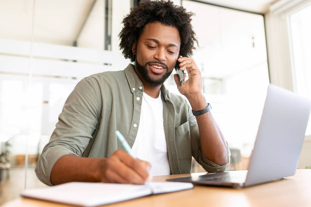 Confident African-American businessman talks phone writing down some notes sitting in the office, friendly male employee speaks on mobile phone with clients, chatting on smartphone in front of laptop - 写真・画像