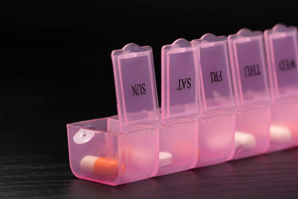 Gros plan d'un pilulier rose rempli, organisateur de pilules, vitamines et additifs alimentaires sur une table noire, macro photographie. Concept de maintien de la santé - Photo, image